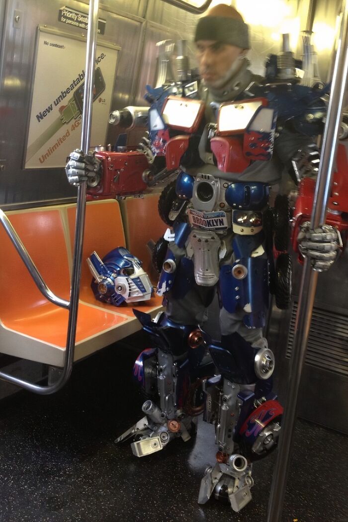 Man in elaborate robot costume holding subway pole inside NYC subway car with empty orange seats and helmet on seat.