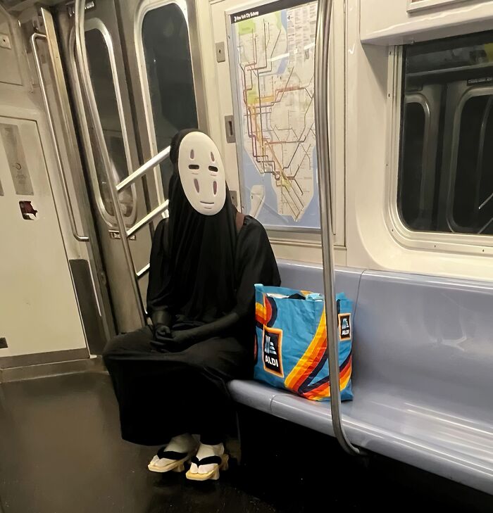 Passenger wearing a No Face costume sits on a NYC subway bench with a colorful Aldi shopping bag beside them.