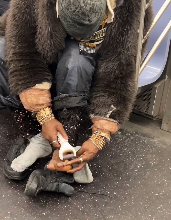 Person on NYC subway with layered bracelets and fur coat, using a handheld tool while sitting on the floor.