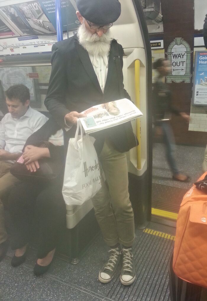 Elderly man with white beard reading newspaper on NYC subway, holding shopping bag, standing near train doors.