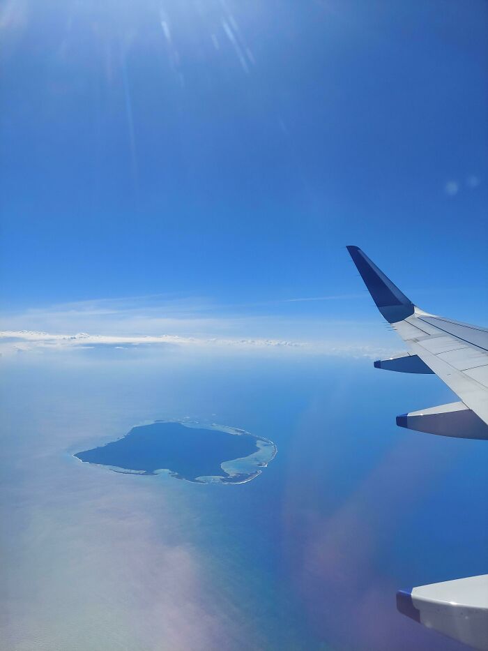 Aerial view from airplane window showing an island surrounded by blue ocean under a clear sky, proving reality is incredible.