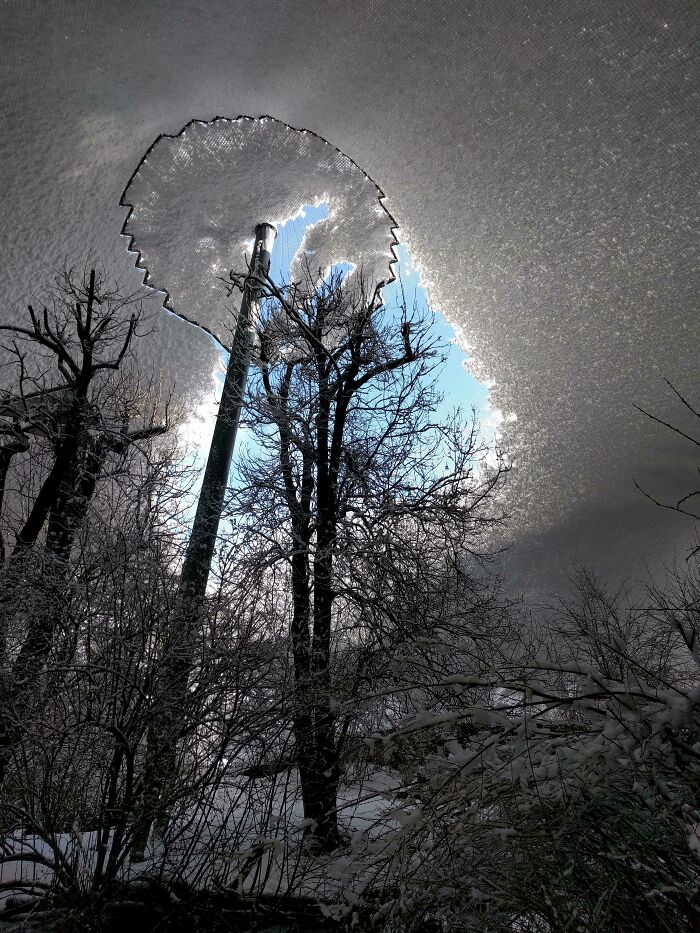 Unreal photo of a snow-covered landscape with a circular hole revealing blue sky above trees and a lamppost.