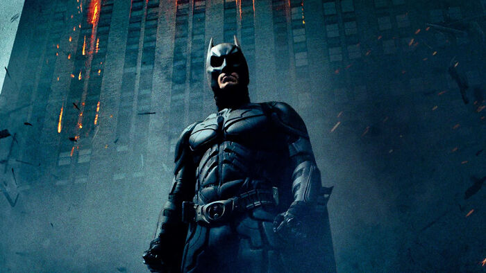 Batman in a dark armored suit stands in front of a burning building scene, highlighting Heat 2 and DC showdown themes.