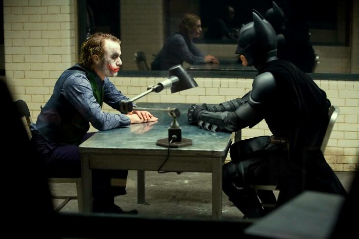 Batman and Joker facing off across a table in a dimly lit interrogation room, highlighting DC showdown tension.