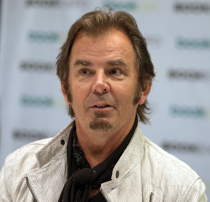 Jonathan Cain speaking at an event, wearing a white jacket and black scarf, highlighting his bio and career achievements.