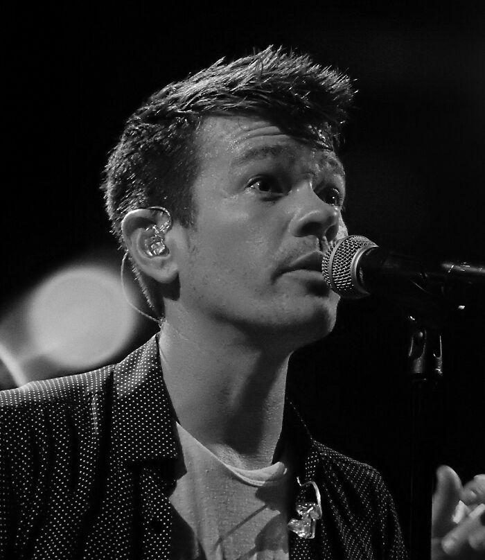 Nate Ruess singing into a microphone on stage, wearing an earpiece and a dark patterned shirt in a black and white photo.