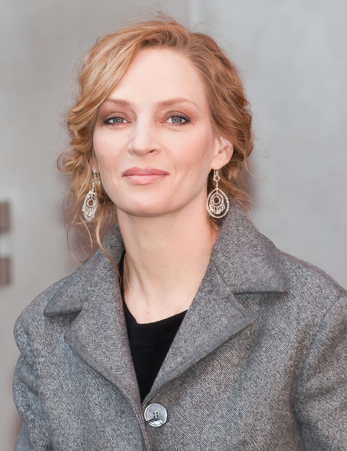 Blonde woman wearing silver earrings and a gray coat, smiling softly in a neutral indoor setting.