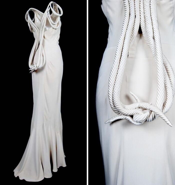 White historical garment with large rope detail draped over a long elegant dress on a black background.