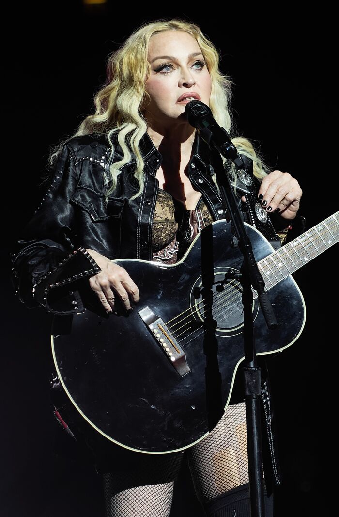 Female musician with blonde hair performing with a black acoustic guitar on stage during a concert.