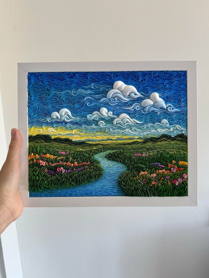 Hand holding a framed colorful DIY landscape artwork showing a river, grassy fields, and clouds under a blue sky.