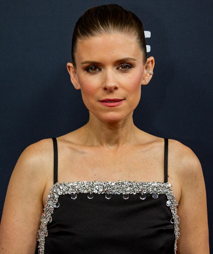 Kate Mara wearing a black dress with silver embellishments, posing against a dark background for a portrait photo.