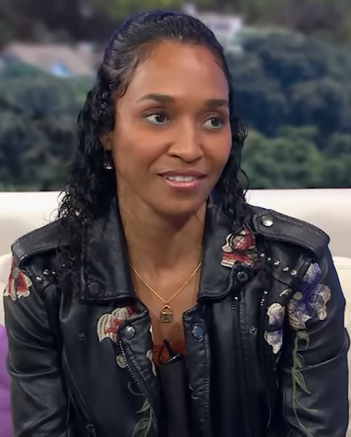 Rozonda Thomas seated, wearing a black floral leather jacket, smiling during an outdoor interview setting.