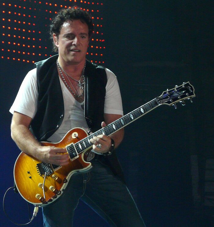 Neal Schon playing electric guitar on stage wearing a white shirt and black vest during a live performance.