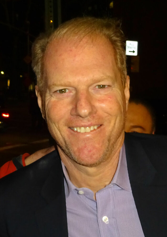 Noah Emmerich smiling outdoors at night, wearing a light purple shirt and dark blazer in a casual setting.