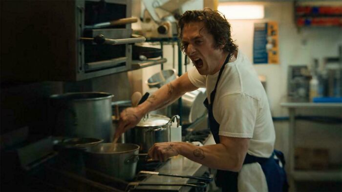 A male chef with curly hair yells while stirring a pot in a chaotic kitchen, depicting hilarious movie details.