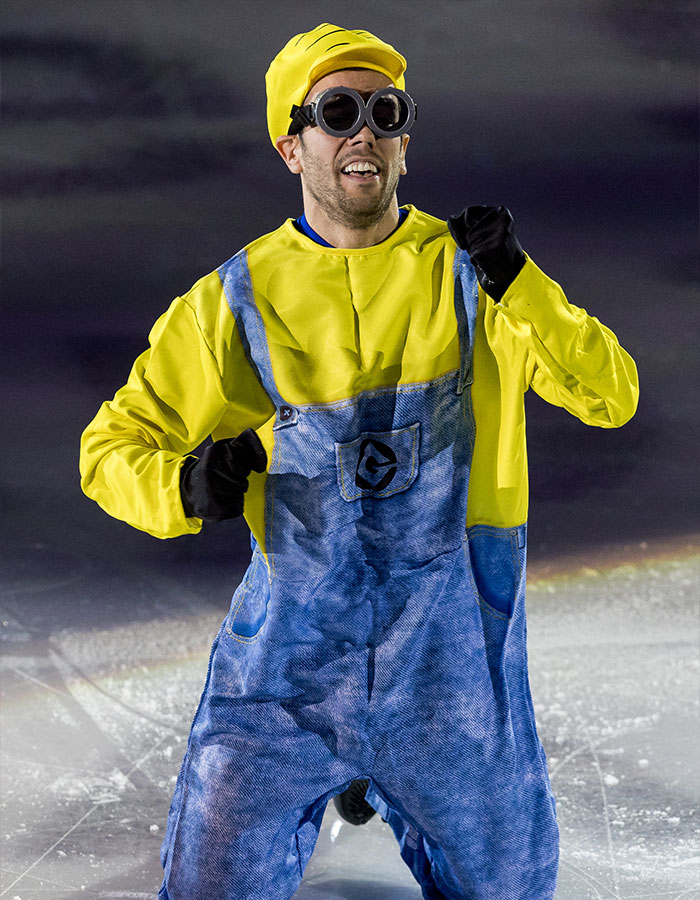 Figure skater dressed as a minion wearing goggles and yellow costume performing on ice at Winter Games event.