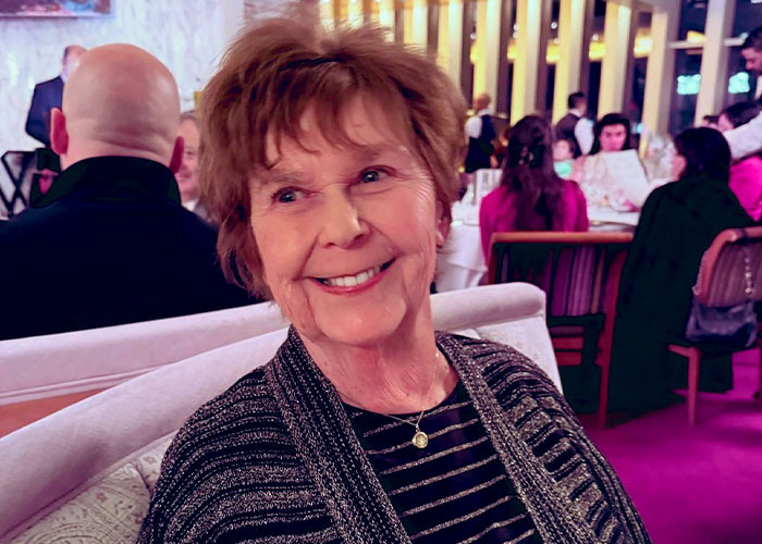 Elderly woman smiling at a social gathering, with blurred people and warm lights in the background.