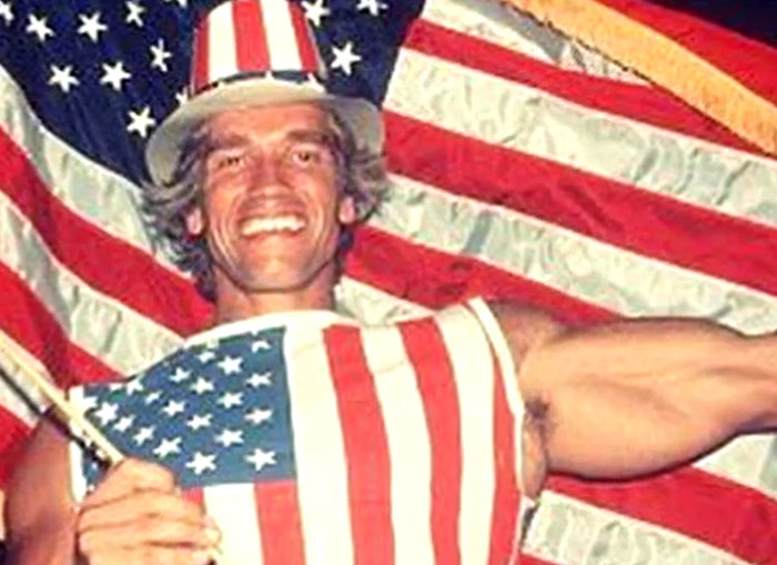 Man wearing patriotic American flag outfit and hat, holding a small flag in front of large US flags, rare historical photo.