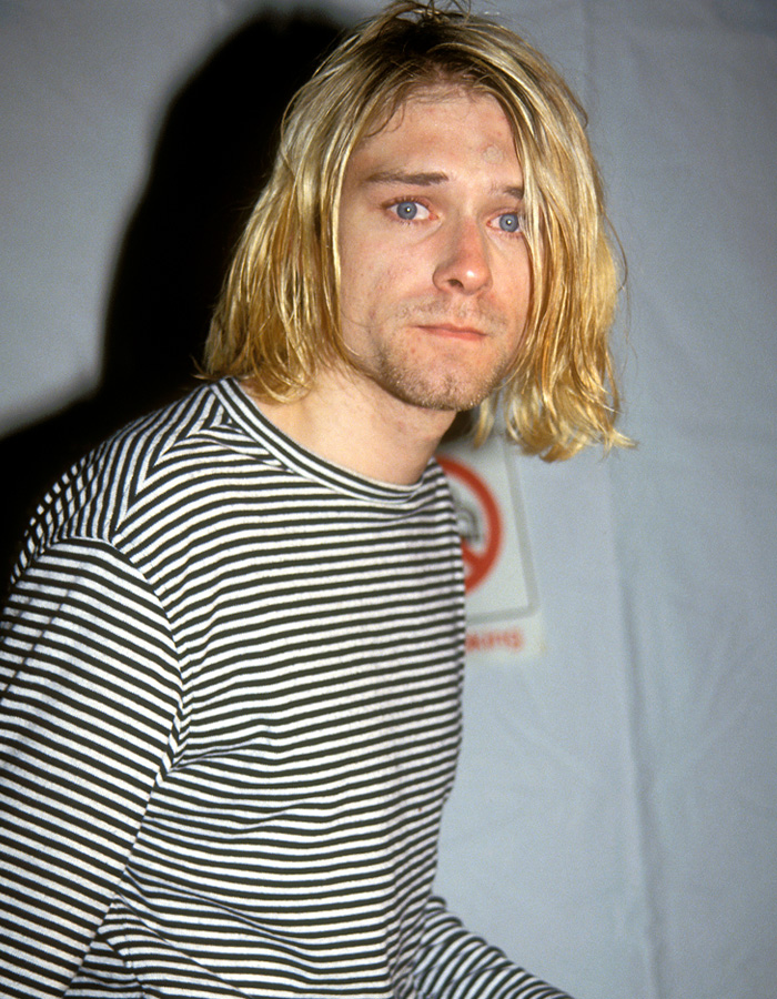 Kurt Cobain in a striped shirt with long blond hair, captured in a candid moment with a neutral expression.
