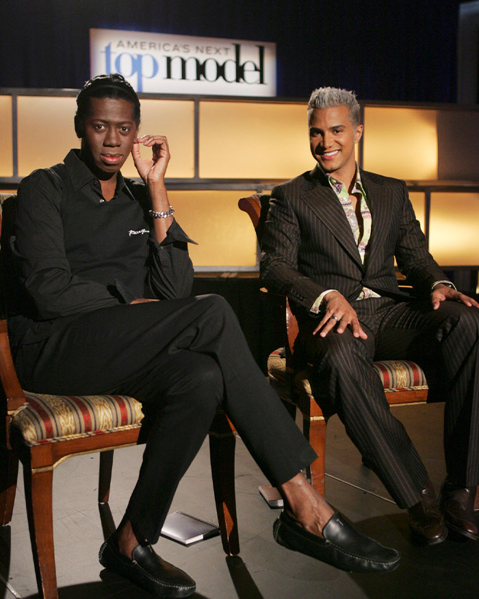ANTM star J Alexander sitting with a male colleague on set of Americas Next Top Model in formal attire