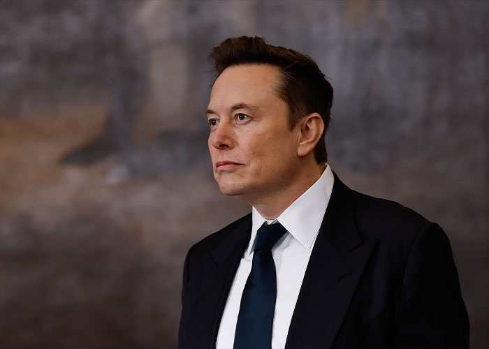 Elon Musk in a dark suit and navy tie looking thoughtfully to the side against a blurred gray background.