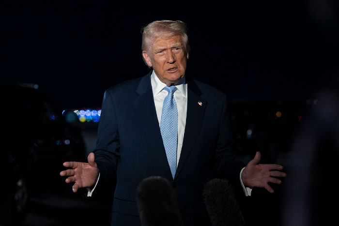 Donald Trump speaking at night, dressed in a suit and tie, amid media microphones during a public event.