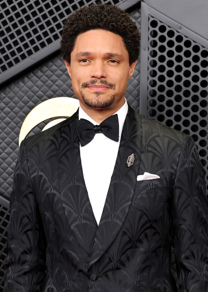 Trevor Noah wearing a patterned black tuxedo with a bow tie, posing against a geometric black and gold backdrop. Trevor Noah wearing a patterned black tuxedo with a bow tie, posing against a geometric black and gold backdrop.