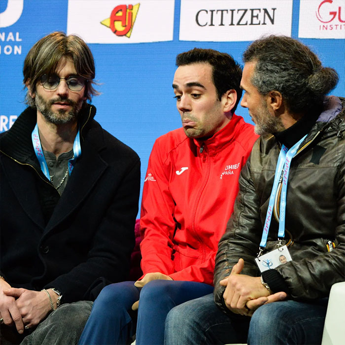 Three men seated talking at an event, with one wearing a red jacket, discussing Olympic figure skater's music banned Winter Games.