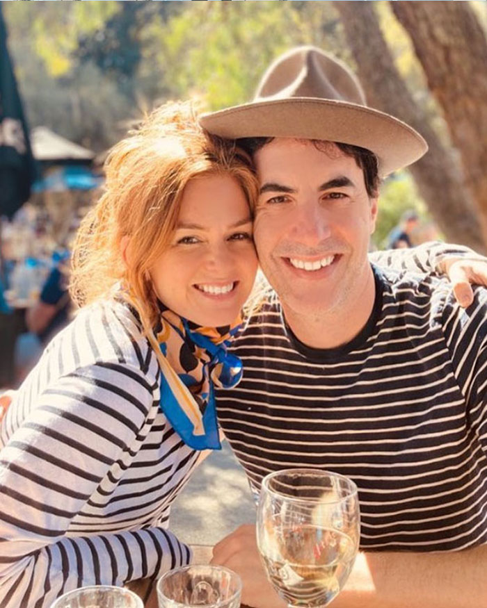 Sacha Baron Cohen and Isla Fisher smiling together outdoors, wearing striped shirts and enjoying a sunny day.