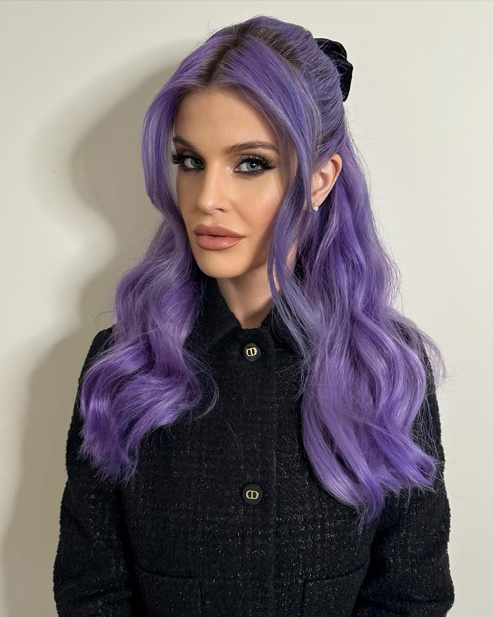Kelly Osbourne with purple hair and stylish makeup wearing a black textured jacket in a neutral indoor setting