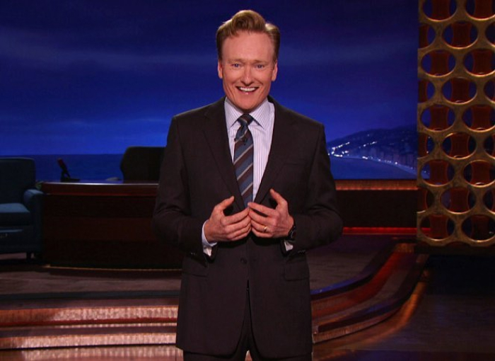Conan O&rsquo;Brien standing on a stage in a dark suit and striped tie, smiling while hosting a late-night show.