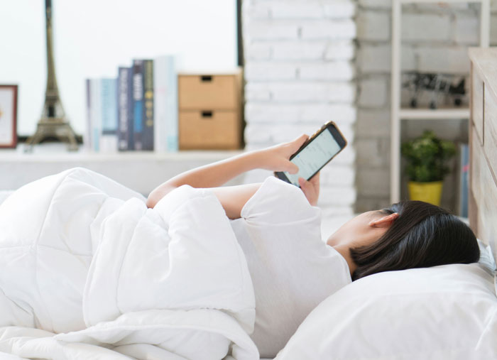 Woman uses phone in bed while waking up boyfriend in a cozy bedroom, showing normal couple morning routine.