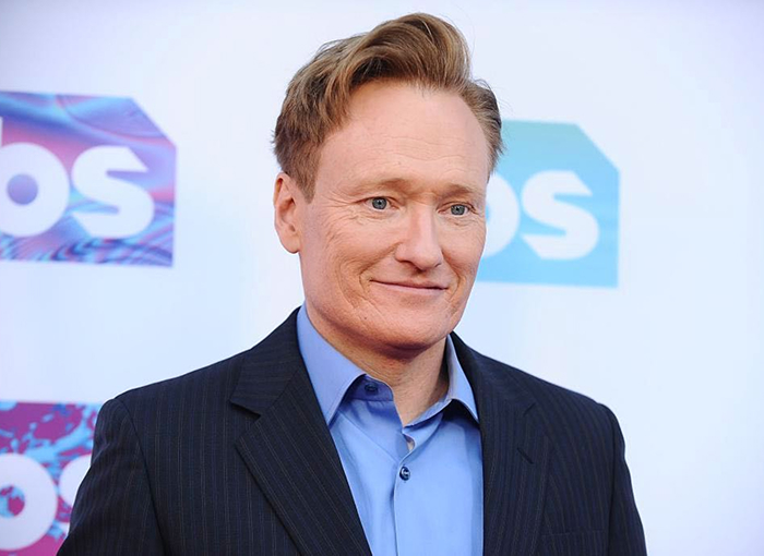 Conan O&rsquo;Brien at a formal event, smiling with a blue shirt and dark suit, linked to hosting Oscars 2026 news.