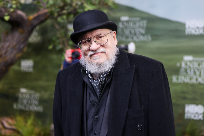 George R.R. Martin at A Knight Of The Seven Kingdoms premiere wearing black coat and hat outdoors with green background. George R.R. Martin at A Knight Of The Seven Kingdoms premiere wearing black coat and hat outdoors with green background.