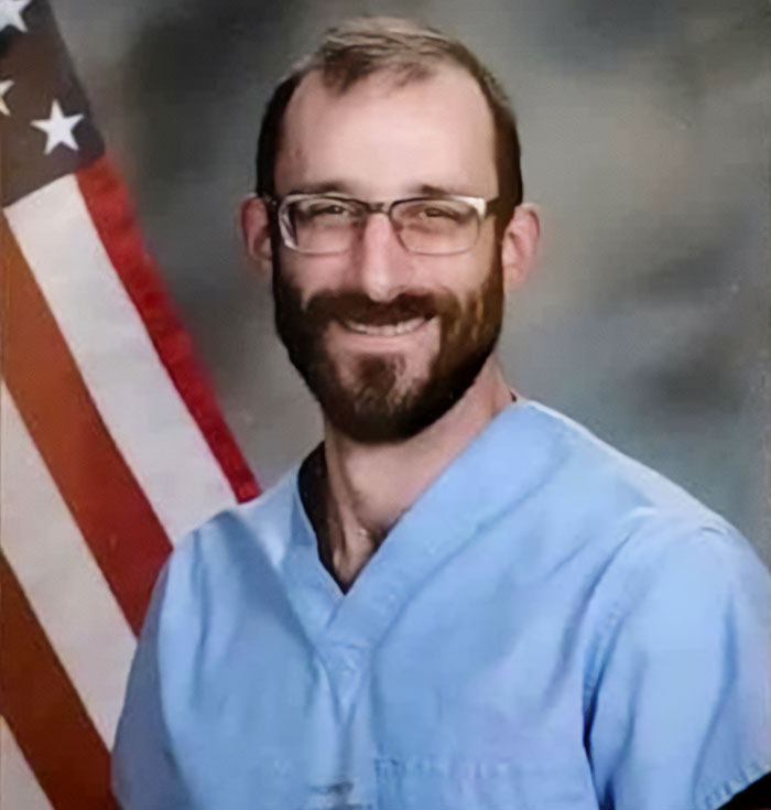 Man in blue medical scrubs and glasses posing in front of American flag, representing medical examiner in Alex Pretti case update