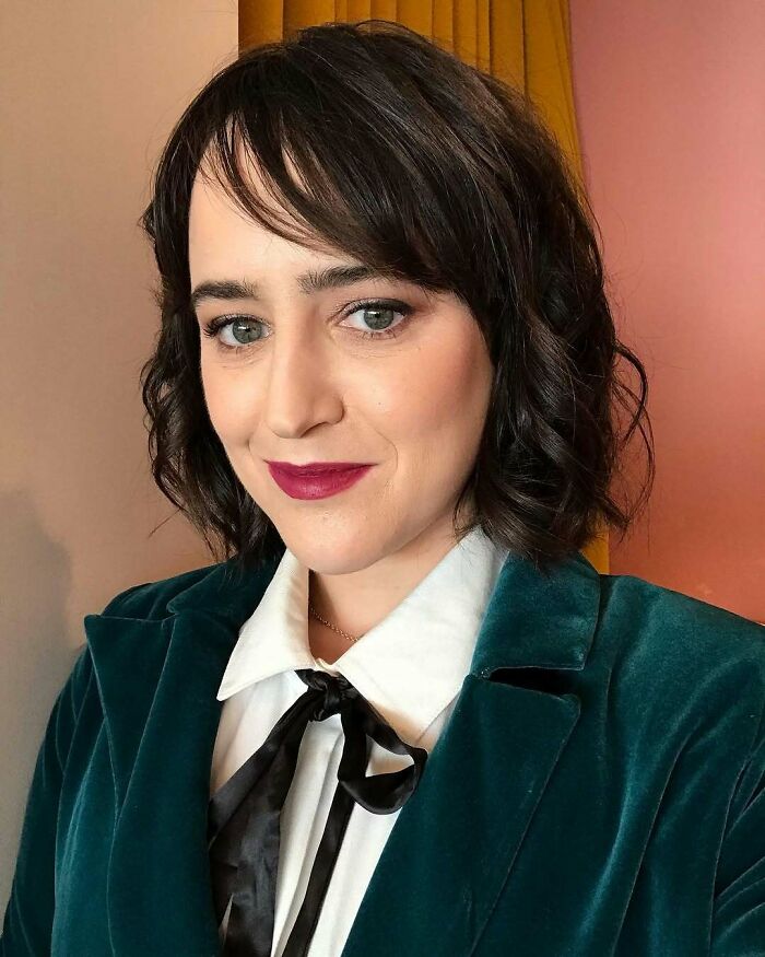 Woman with short dark hair and red lipstick wearing a teal velvet blazer and white shirt, representing iconic actors in Hollywood.
