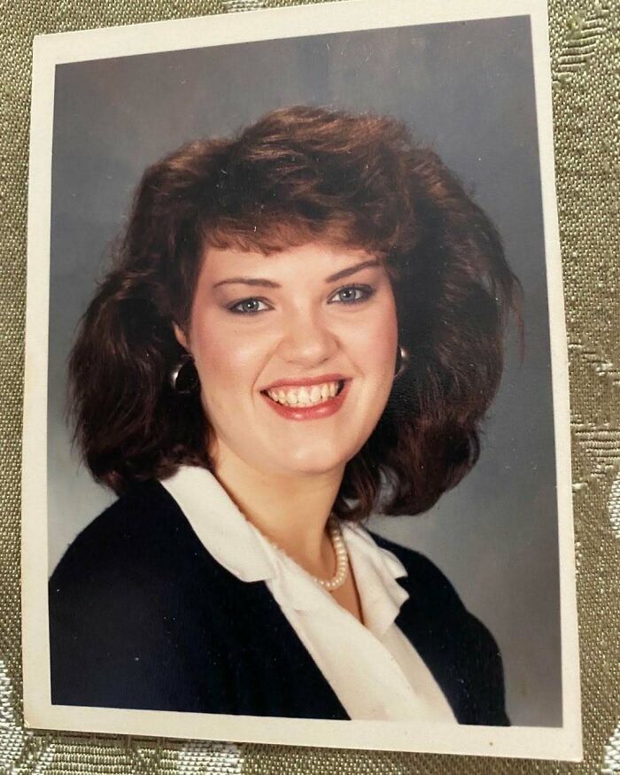 Woman with an iconic 80s hairdo featuring voluminous, teased hair, wearing hoop earrings and a pearl necklace, smiling.