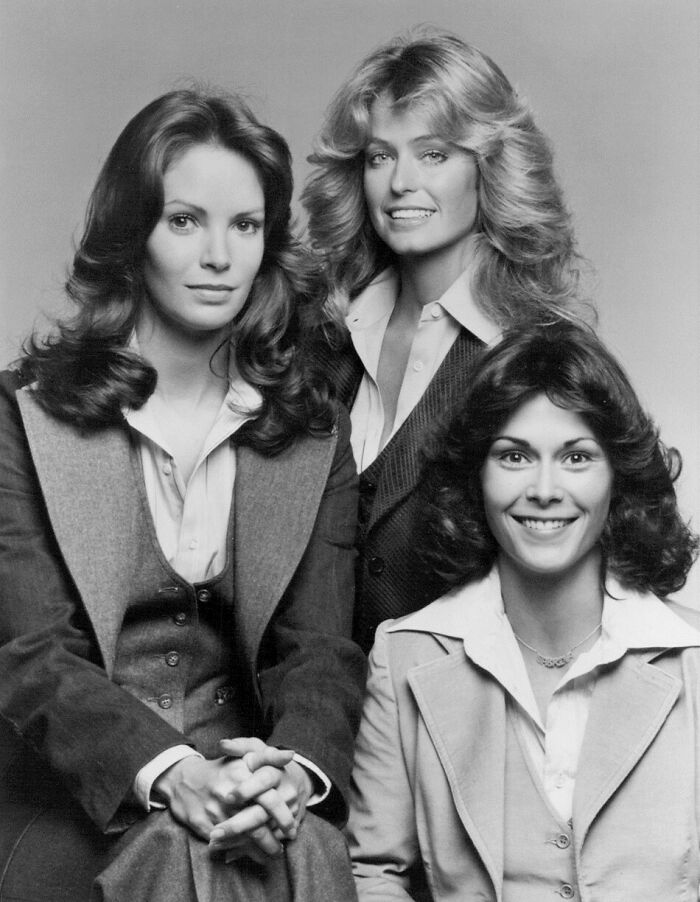 Three women with iconic 80s hairdos including feathered and voluminous styles, dressed in vintage suits and smiling for a studio portrait.