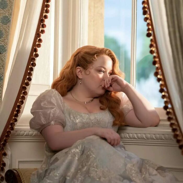 Woman in historic dress sitting thoughtfully by a window, captured for Bridgerton memes fans of the show.