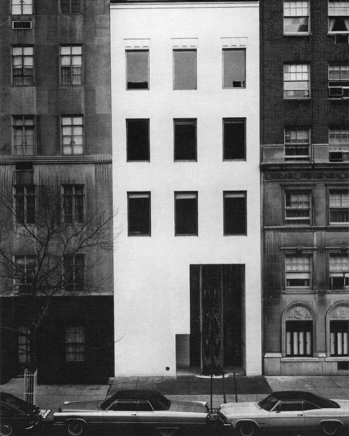Minimalist white architectural structure with rectangular windows, surrounded by classic urban buildings showcasing creative architectural design.