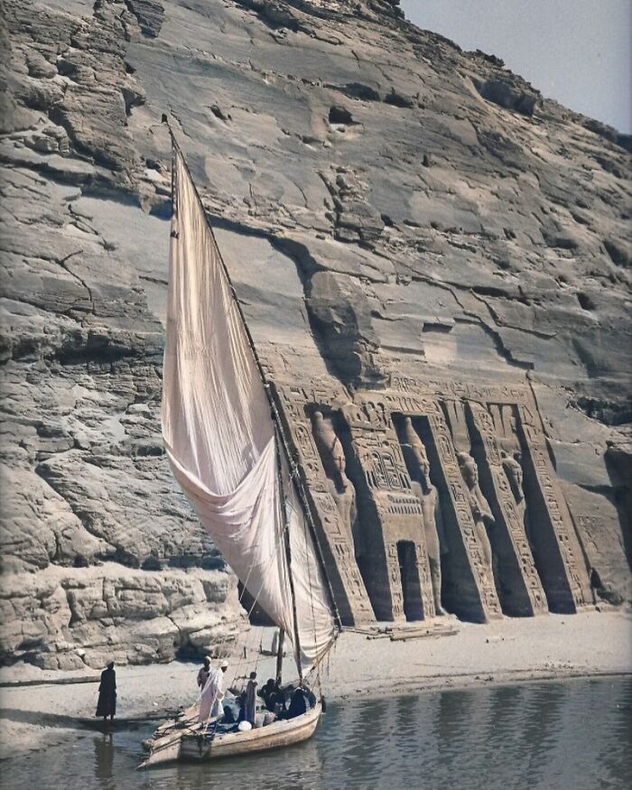 Ancient architectural structures carved into rock face beside a river with a traditional sailboat and people nearby.