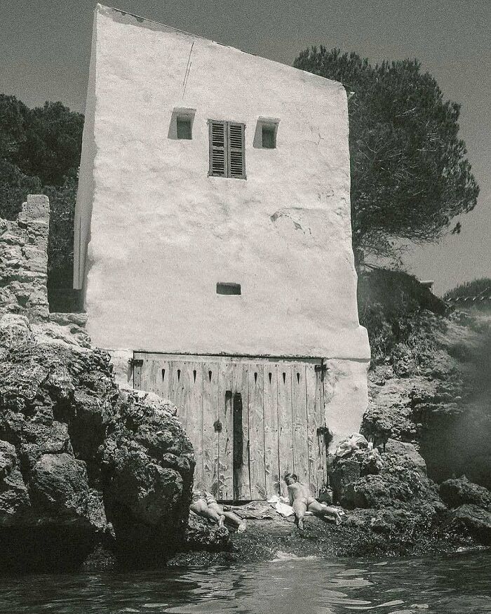 Old whitewashed architectural structure by rocky shore with people sunbathing, showcasing humanity's creativity in design.