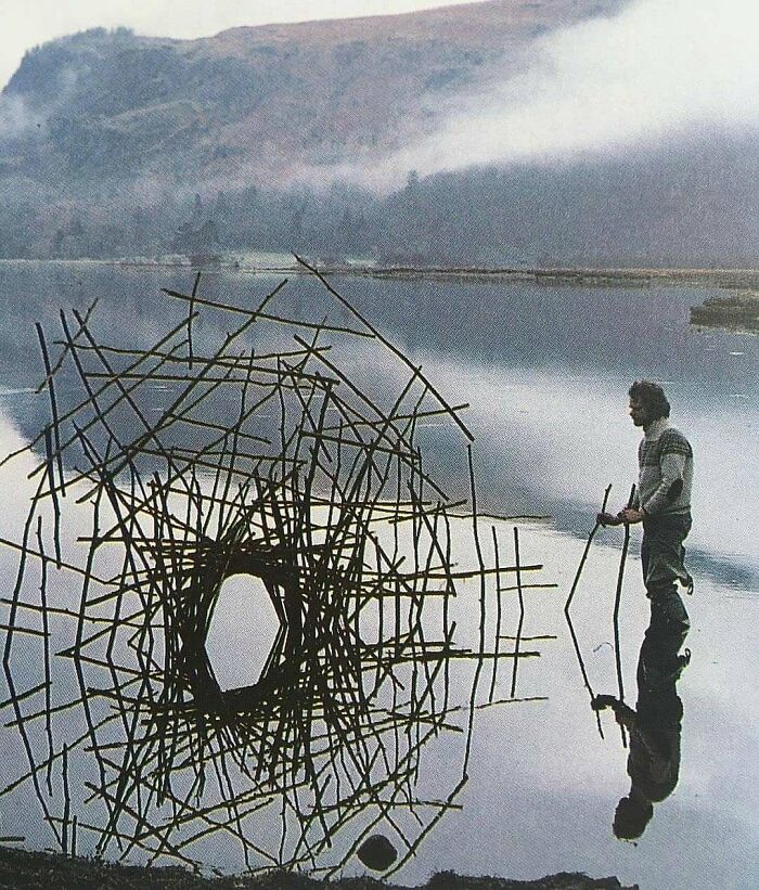 Man standing near an intricate architectural structure made of sticks reflected in a calm body of water showcasing creativity.