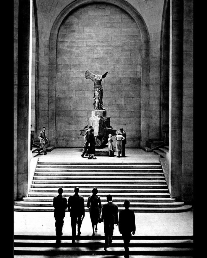 Visitors ascending grand stairs inside an architectural structure featuring a large classical statue on a pedestal.