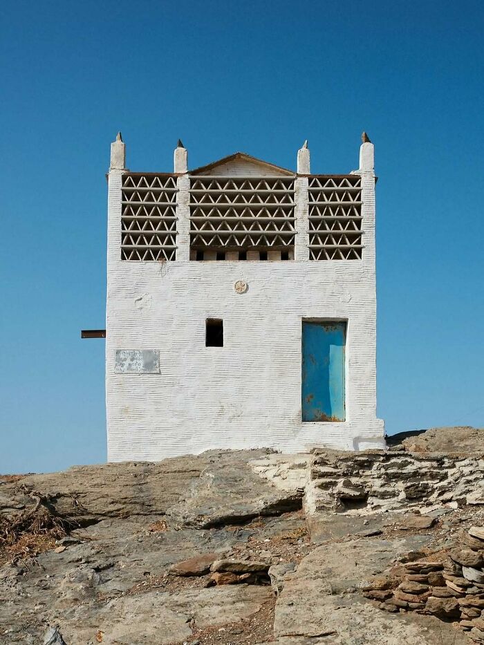 White architectural structure with unique triangular patterns on top, standing on rocky terrain under clear blue sky.