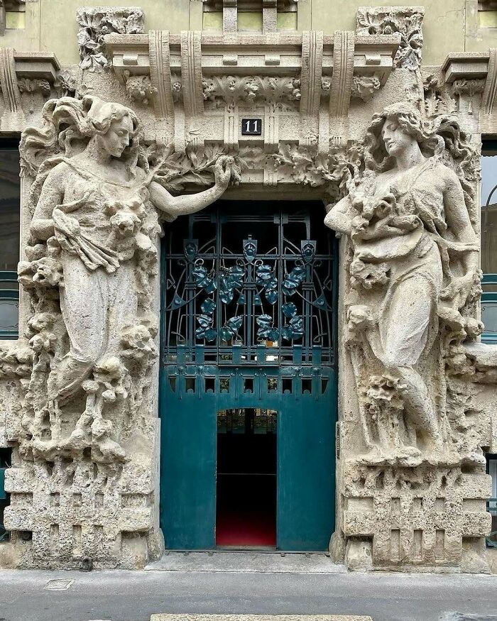 Ornate architectural structure with detailed stone sculptures of two figures flanking an intricate wrought iron doorway.
