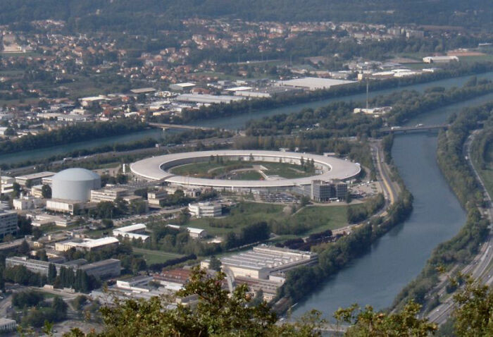 Aerial view of a large circular building complex near a river, surrounded by urban and green areas.