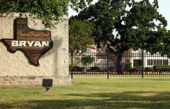 Federal Prison Camp Bryan entrance sign with Texas-shaped wooden plaque, surrounded by greenery and a fence in daylight. Federal Prison Camp Bryan entrance sign with Texas-shaped wooden plaque, surrounded by greenery and a fence in daylight.