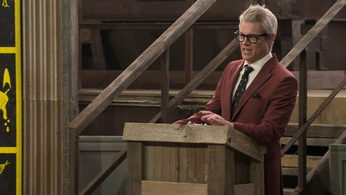 Host standing behind a wooden podium on Fear Factor House of Fear set, preparing for episode 6 release.
