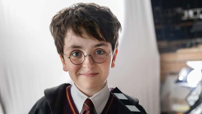 New Draco Malfoy actor smiling with round glasses and Hogwarts robe, discussing difference between Harry Potter series and movies.